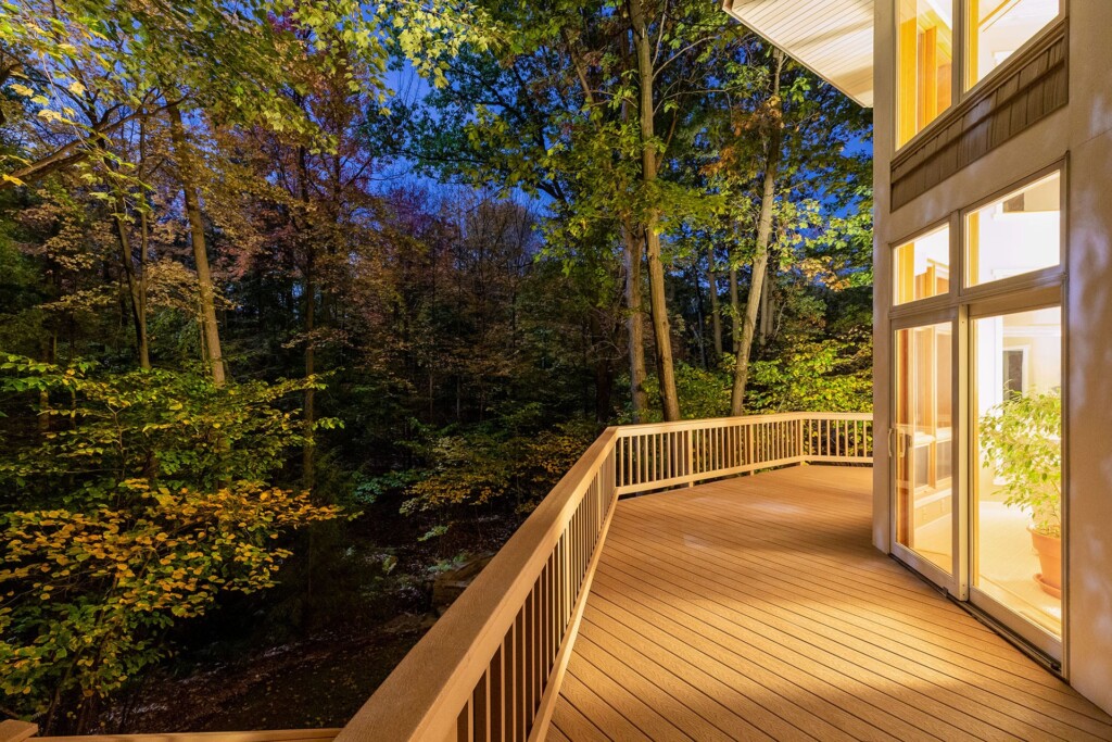 Modern wooden deck overlooking lush forest at dusk with warm indoor lighting.